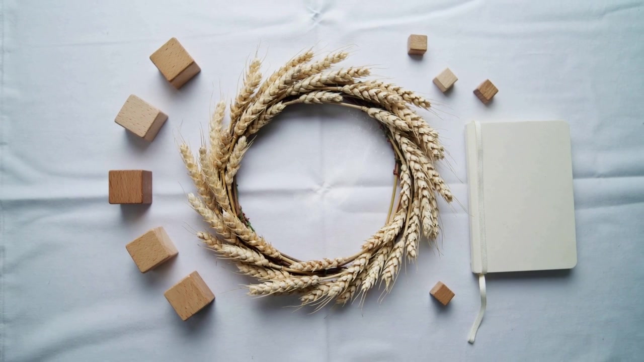 Camera shifting upward, widening view on pale cloth, showing wheat wreath, blocks, white journal