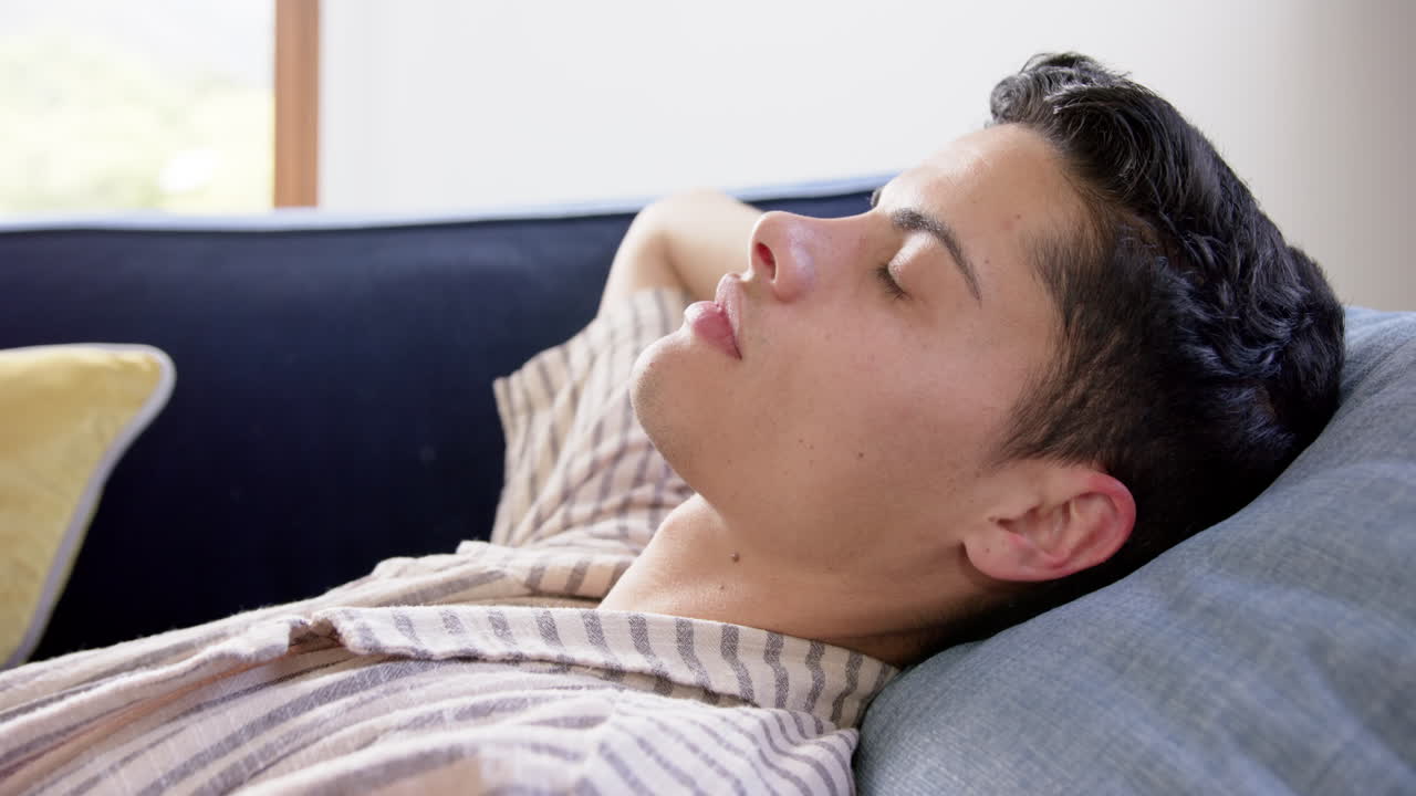 hombre biracial cansado acostado en el sofá descansando con los ojos cerrados, cámara lenta