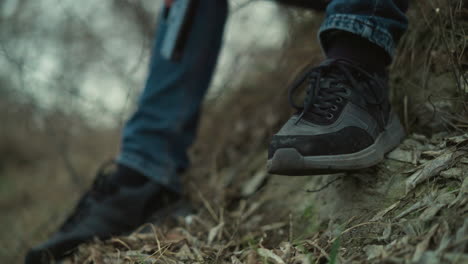 a close view of a person's feet wearing black sneakers tipping his one leg, sitting on a grassy slope, the person is holding a black handgun