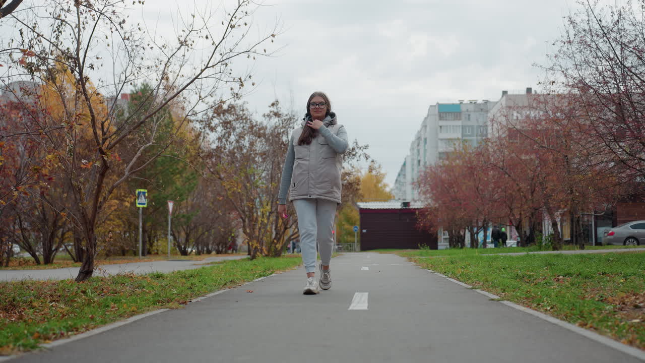 Freelancer walking on tarred road adjusting hair while others walk by in hoodies and car parked in background with autumn trees, residential buildings, and green grass lining