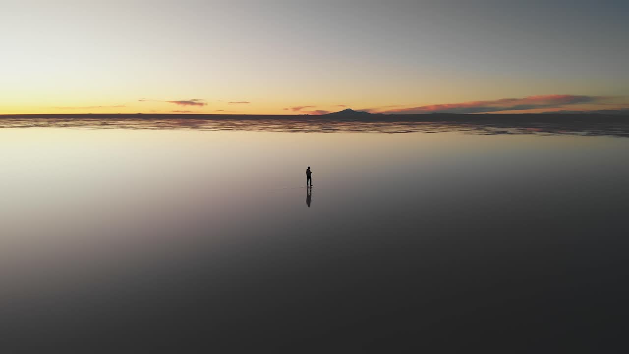 Aerial of a lone figure slowly walking along the mirrored reflection of the world's largest salt flat at dusk in Uyuni Salt Flats , Bolivia