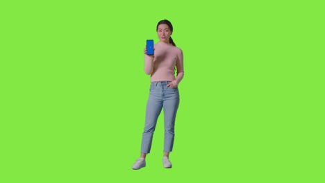 full length studio portrait of smiling woman holding blue screen mobile phone towards camera against green screen