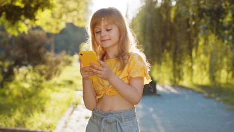 smiling child girl using smartphone typing text answering messages chatting online in city street
