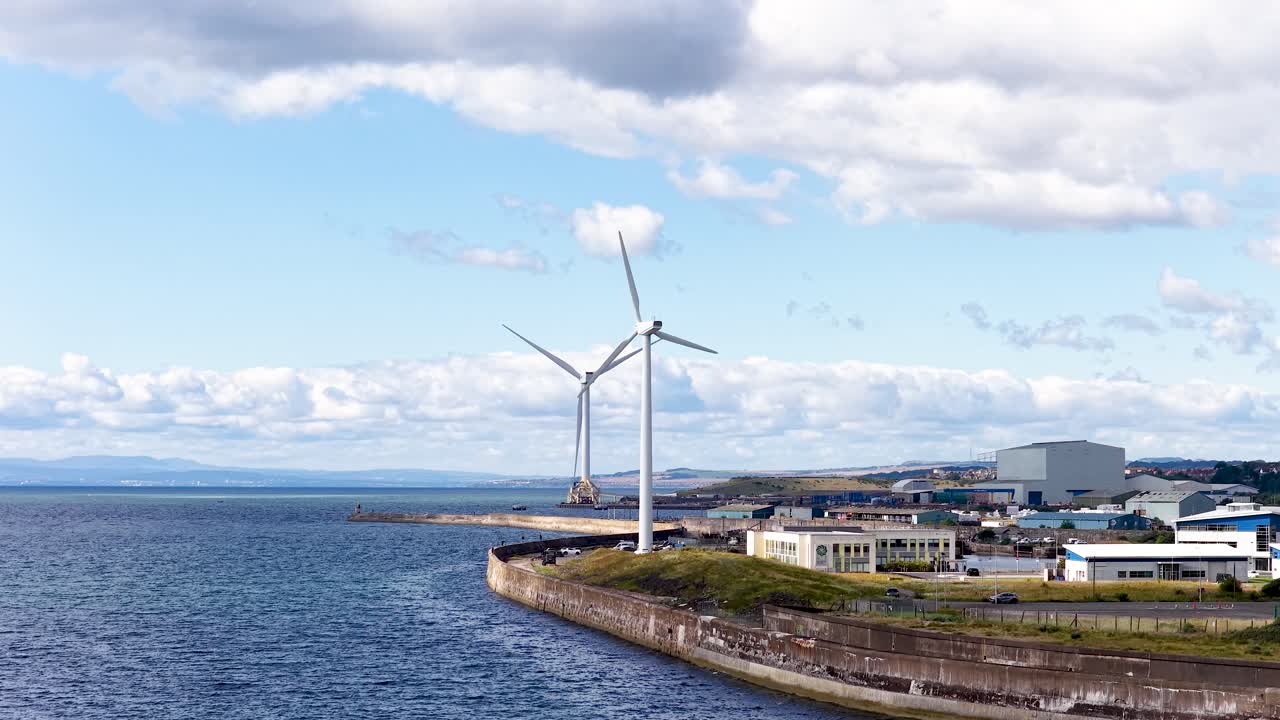 Large wind turbine spins beside water, industrial buildings, and coastline under bright daylight, static camera