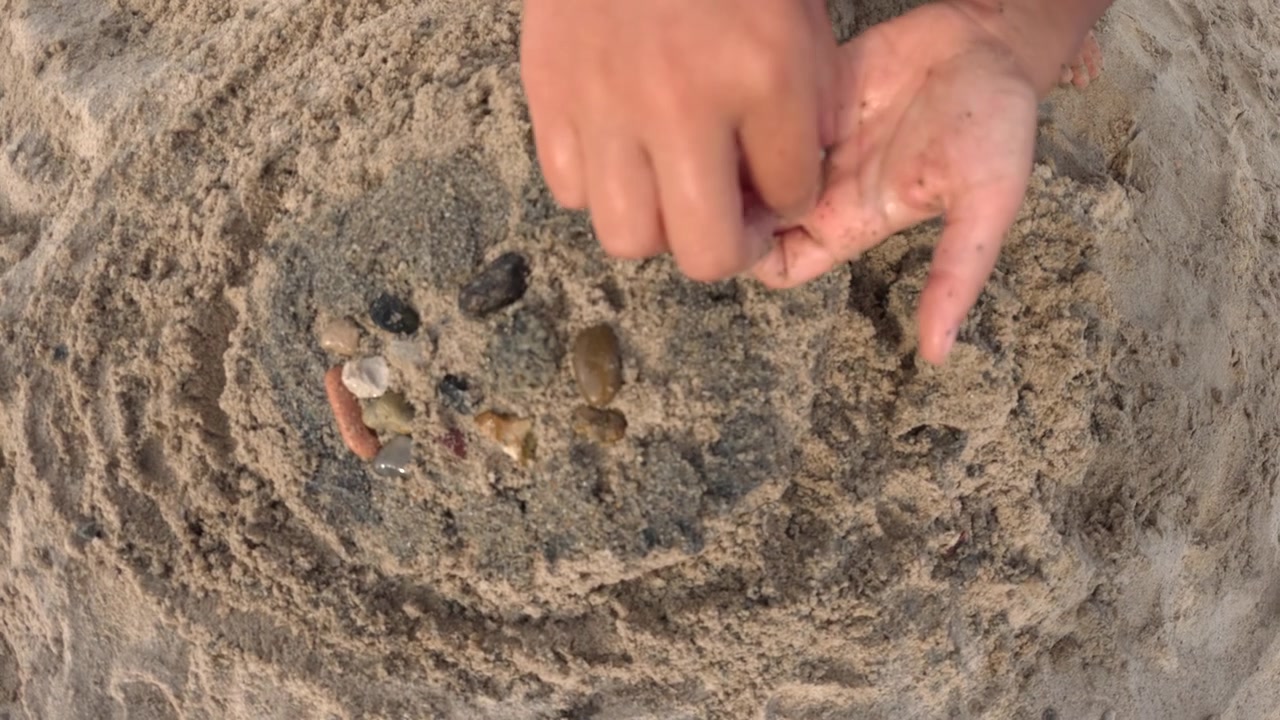 Close-up of a child's hands arranging small colorful pebbles and shells on a sand mound, Golfe-Juan, France