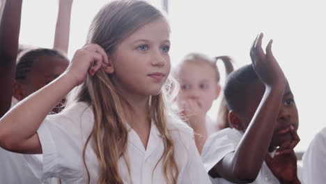 group of elementary pupils wearing uniform sitting on floor raise hands to answer question in class