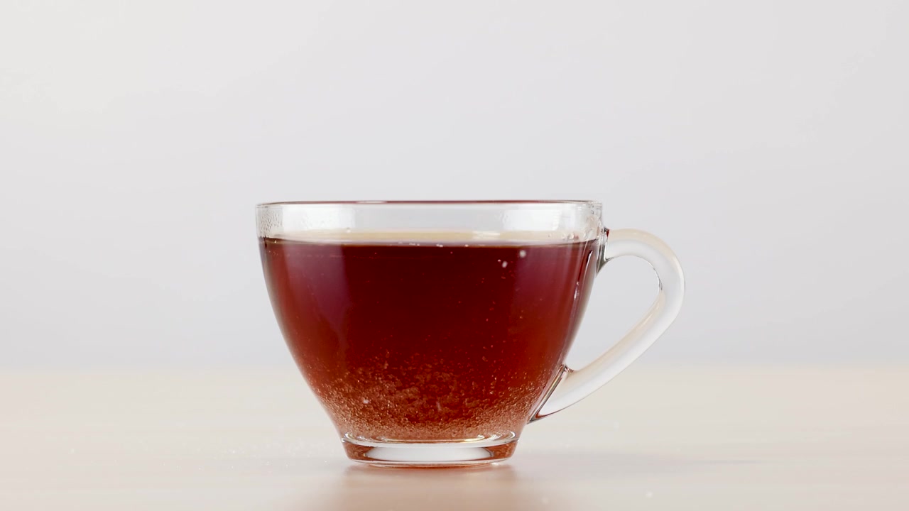 Granulated sugar dissolves at the bottom of a clear glass mug filled with hot tea.