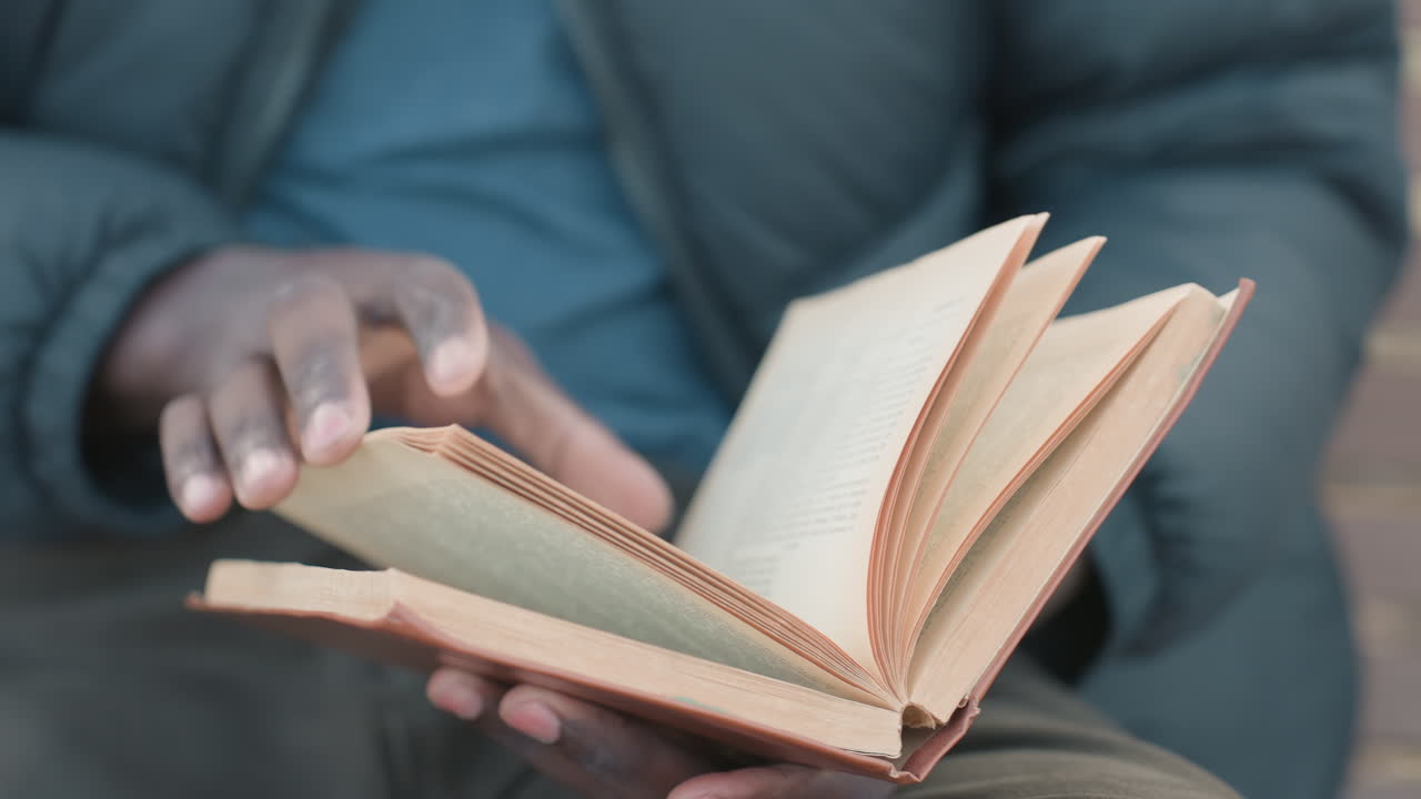 Close up of adult reading novel outdoors, hands holding worn book on lap, pages turning to new chapter under soft daylight with blurred background