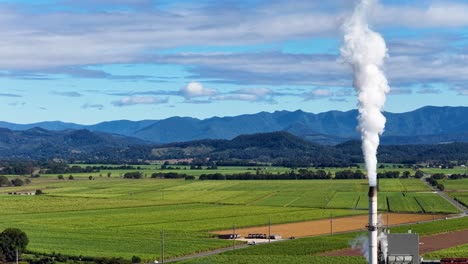 a tall smokestack emits white smoke into a clear sky over expansive green fields, with distant mountains under bright daylight