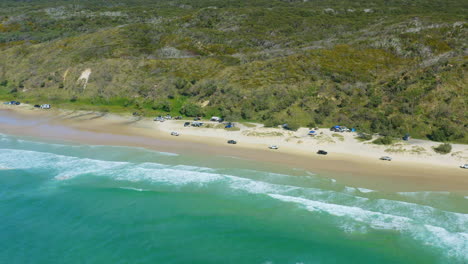 blue ocean waves beside 4wd cars driving along sandy beach shore, 4k aerial drone