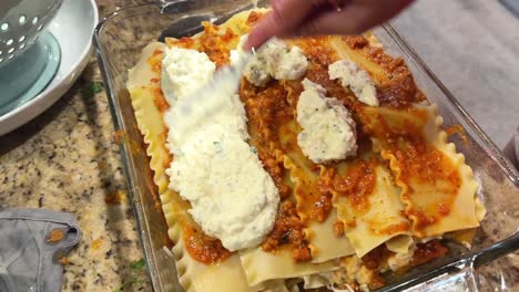 woman making homemade lasagna, focused on a close up of the hand holding a fork spreading ricotta cheese on top layer