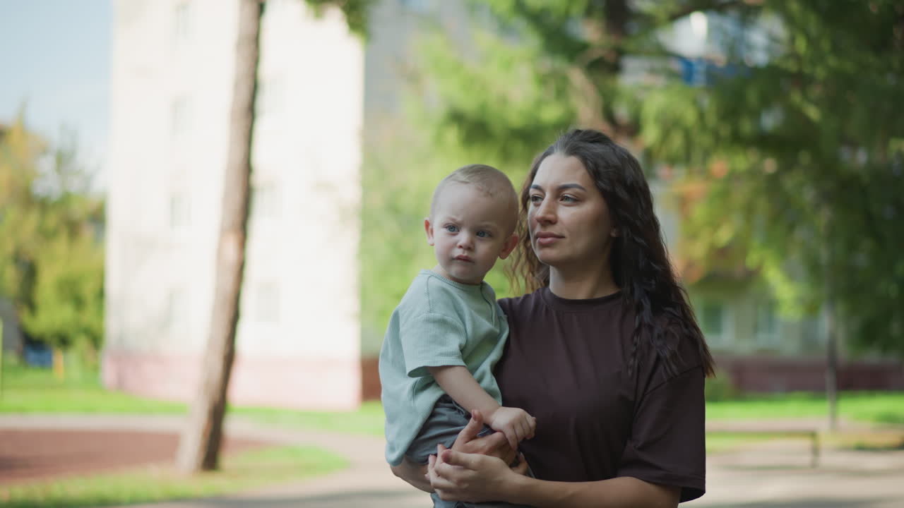 Madre latina che tiene in braccio un bambino piccolo al parco, sguardo contemplativo vicino a un condominio, alberi verdi e luce solare soffusa, tranquilla scena pomeridiana che mostra uno stretto legame genitore-figlio e un umore premuroso e discreto.