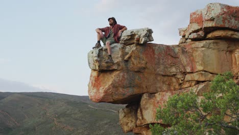 feliz caucásico hombre sobreviviente sentado en el pico de la montaña rocosa en el desierto, disfrutando de la vista