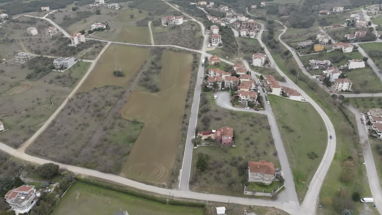High-angle aerial drone shot moves towards picturesque village with scattered houses, winding roads and green fields.
