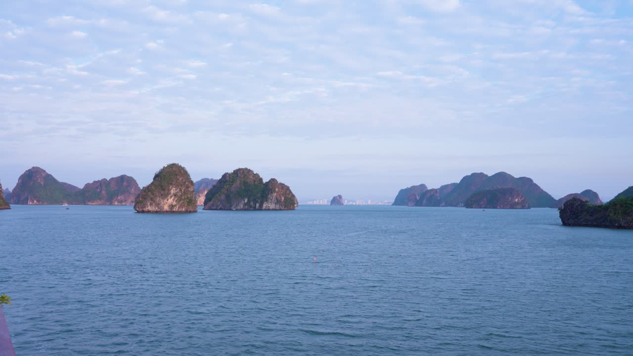 Majestic limestone karsts rise from the tranquil turquoise waters of Ha Long Bay, creating a mesmerizing seascape where ancient rock formations pierce through the misty horizon under a soft blue sky
