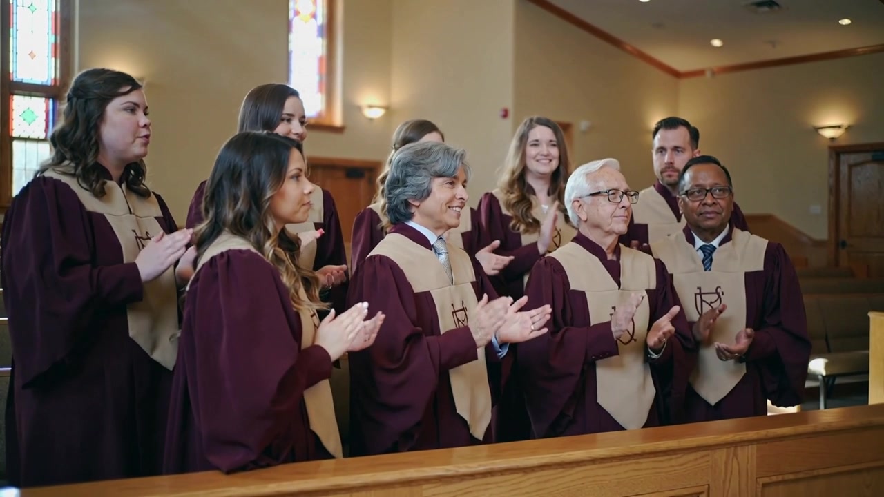 Clapping choir group wearing maroon robes with beige stoles responding to hymn cue in sanctuary