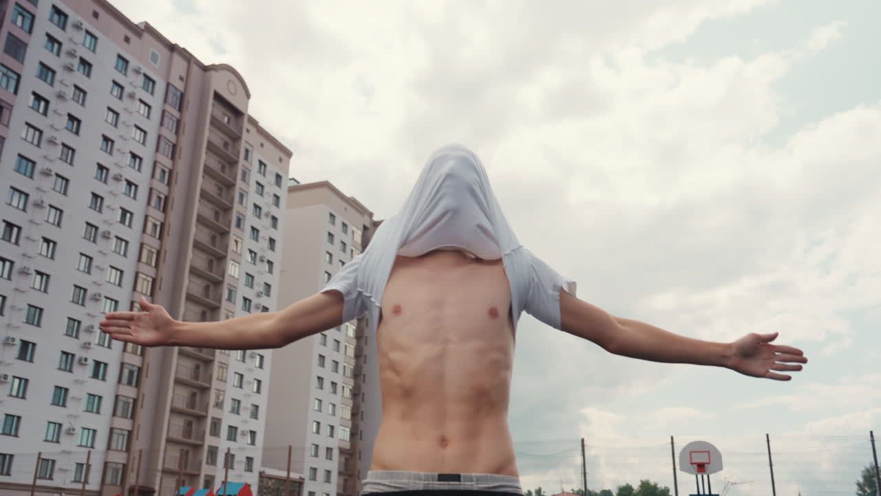 uomo su un campo da basket all'aperto si sfila la maglietta dalla testa scoprendo il busto, allarga le braccia sotto un cielo nuvoloso, inquadrato da alti condomini e un canestro da basket, atmosfera urbana grintosa, espressivo