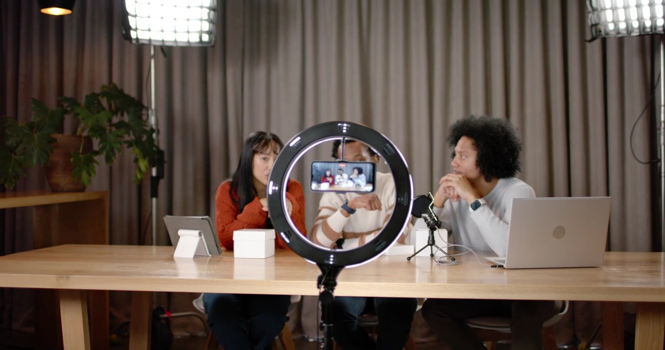 African American co-hosts starting ring light smartphone recording, showing product box at set demo