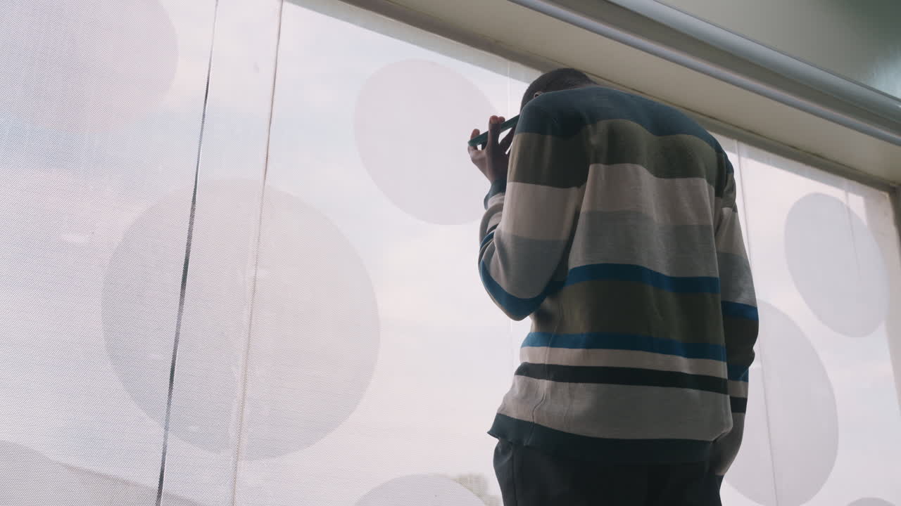 Black man at patterned window waiting. Soft daylight filters through circular curtain pattern, striped sweater, contemplative posture, finger near phone, quiet anticipation for romantic evening
