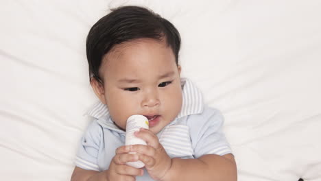 asian baby boy biting on a bottle teether while lying in bed