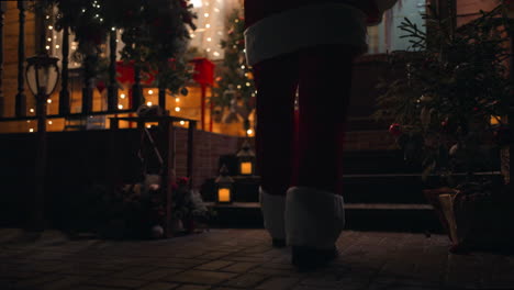 A-view-from-the-back-of-Santa-Claus's-feet-carries-and-delivers-a-gift-in-his-hands-in-winter-and-enters-the-house-with-garlands-and-Christmas-decorations.-In-December-on-Christmas-Eve