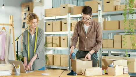 caucasian man working in clothing store packing ordered in parcels while caucasian woman typing on laptop online