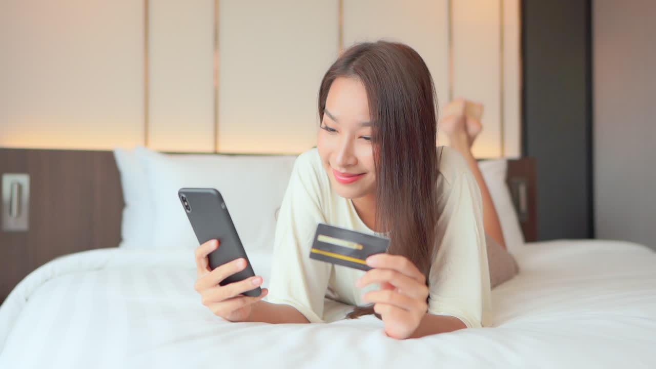 一位年轻女性躺在床上，专注于智能手机上的信用卡信息输入