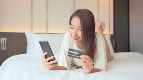 a young woman lying on a bed focuses on her smartphone and credit card input