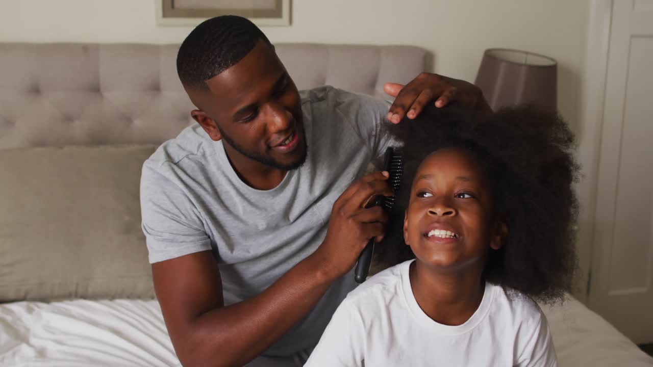 African american father brushing his daughters hair while sitting on the bed at home