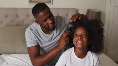 african american father brushing his daughters hair while sitting on the bed at home