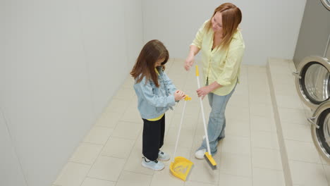 Aerial view shows cheerful mother sweeping tiled laundromat floor while toddler helper holds dustpan, both talking and smiling during cleaning routine beside stainless washers, yellow tools