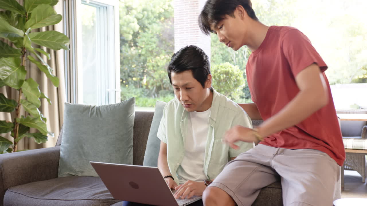 Asian father and teenage son using laptop together in a bright living room