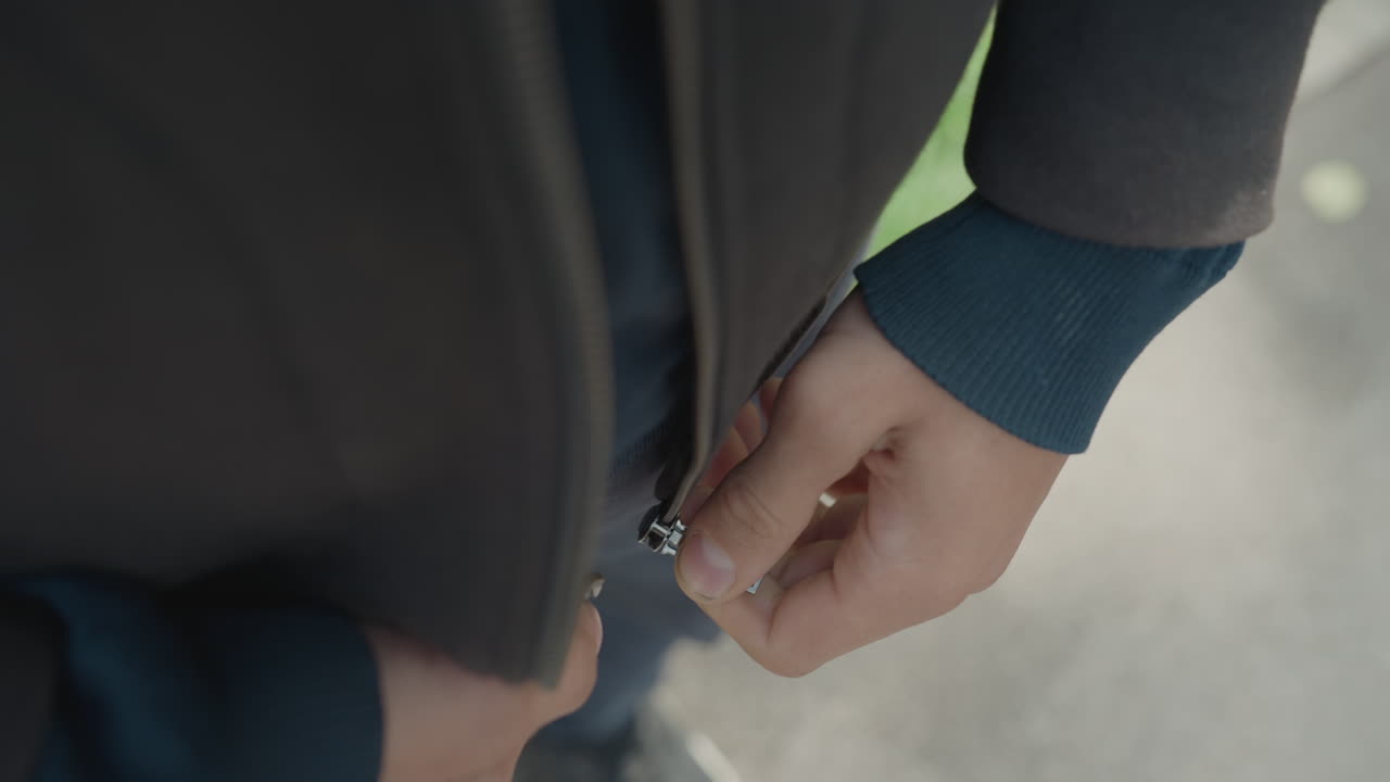 Gros plan sur un homme blanc qui ferme la fermeture éclair de sa veste, les mains tendues lors de la répétition en coulisses, l’attention portée sur le pouce et l’ongle, texture du bord-côte tricoté, mouvement subtil, plan de détail cinématographique intime avec une faible profondeur de champ