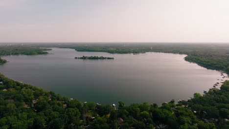 a wide drone shot captures the serene beauty of a large lake surrounded by trees, with a small wooded island nestled in the middle of the calm waters