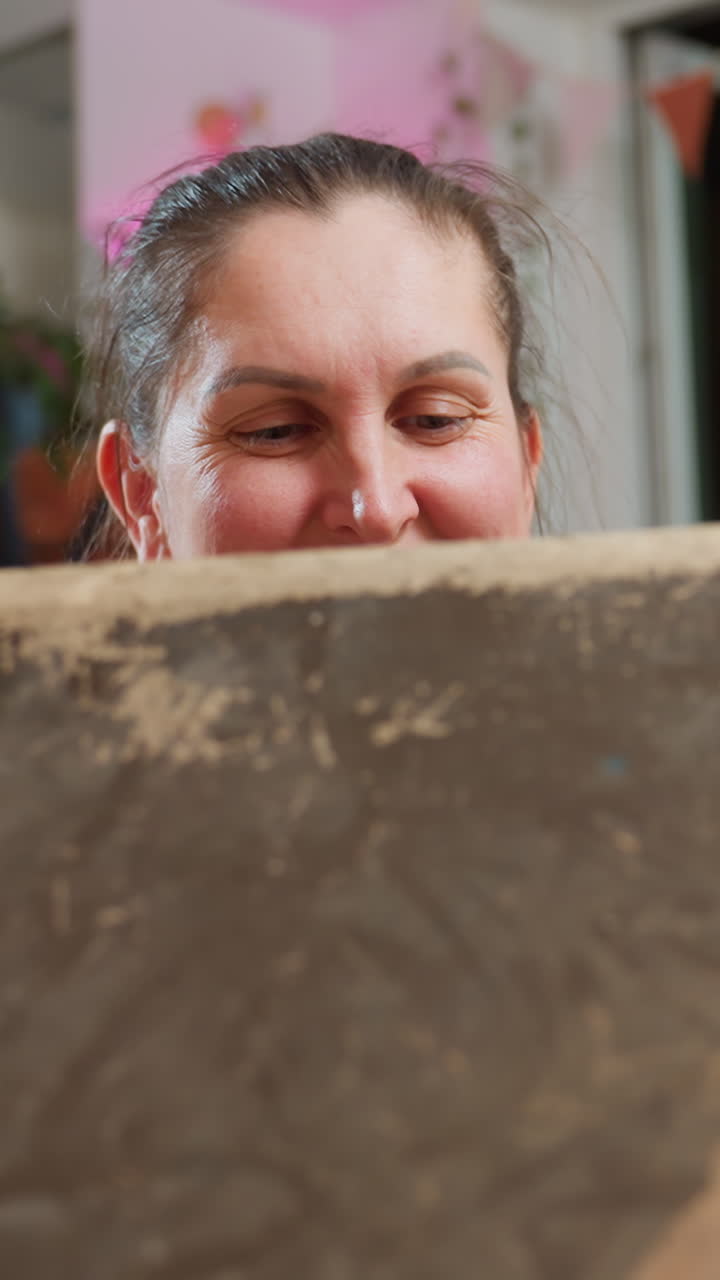 primer plano de una mujer caucásica inspeccionando un lienzo de arcilla, iluminación de estudio acogedora, sonriendo con expresión concentrada mientras sujeta un marco texturizado desgastado, plantas y guirnaldas de banderines al fondo, artesanía casera delicada