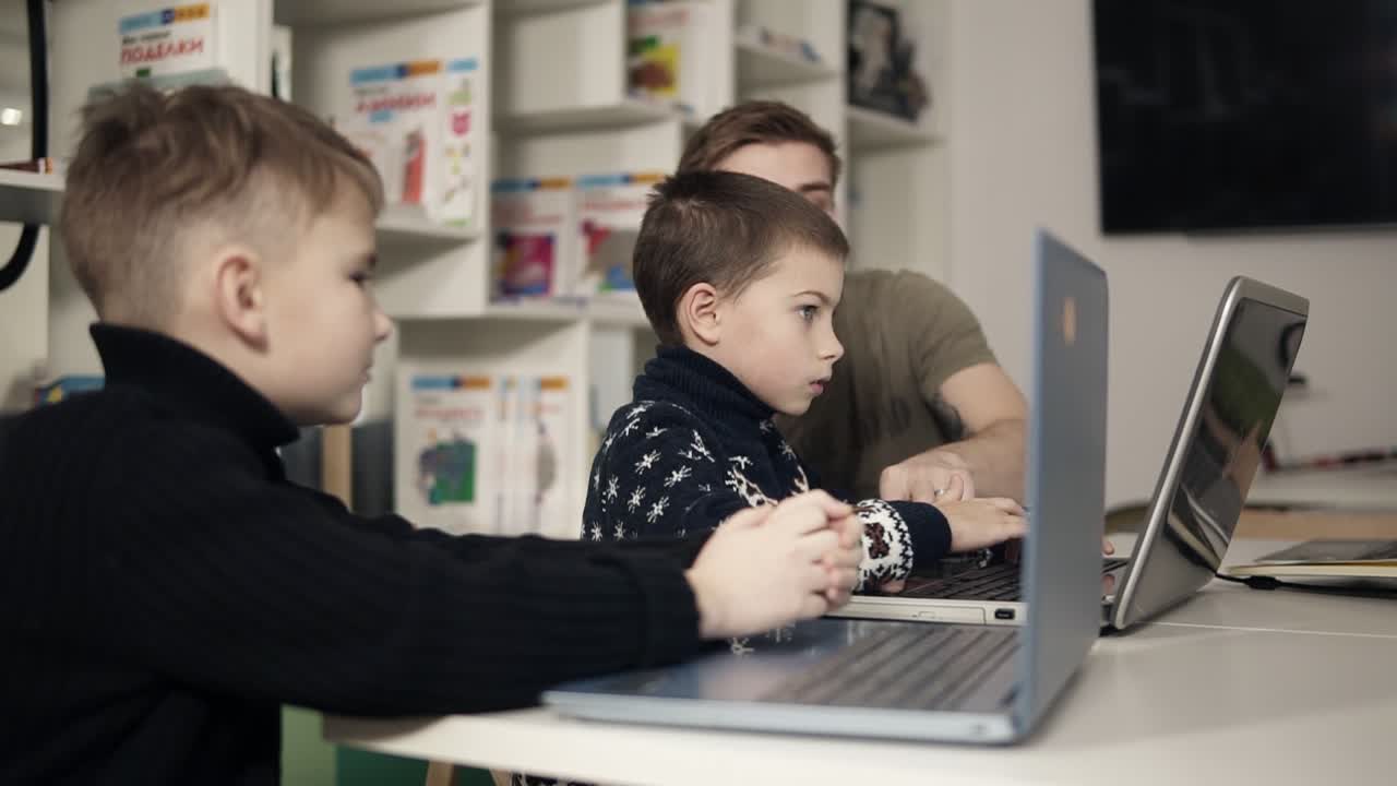 Two young boys are learning how to use laptops with help and guidance of young programmer teacher sitting beside them in a classroom.