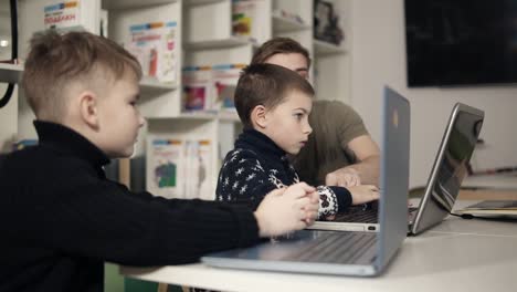 two young boys are learning how to use laptops with help and guidance of young programmer teacher sitting beside them in a classroom.