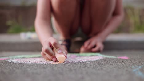 handheld close up front shot of a girl child sitting on the sidewalk drawing a mushroom on the street asphalt pavement with orange colourful chalk in glowing evening light