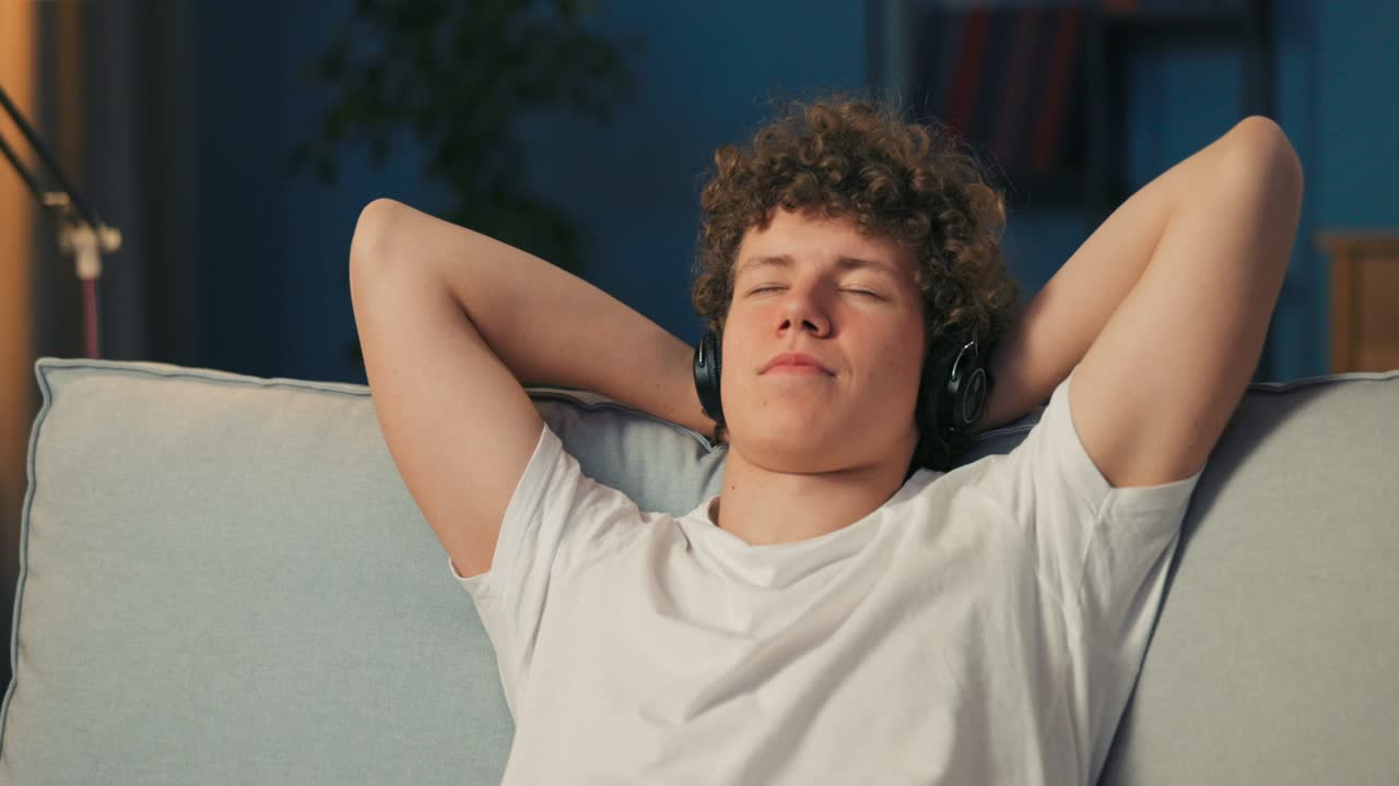 The man is resting on the sofa, hands behind head, listening to music, podcast on wireless headphones on ears, eyes closed, listening to the song, immersed in dreams, falling asleep