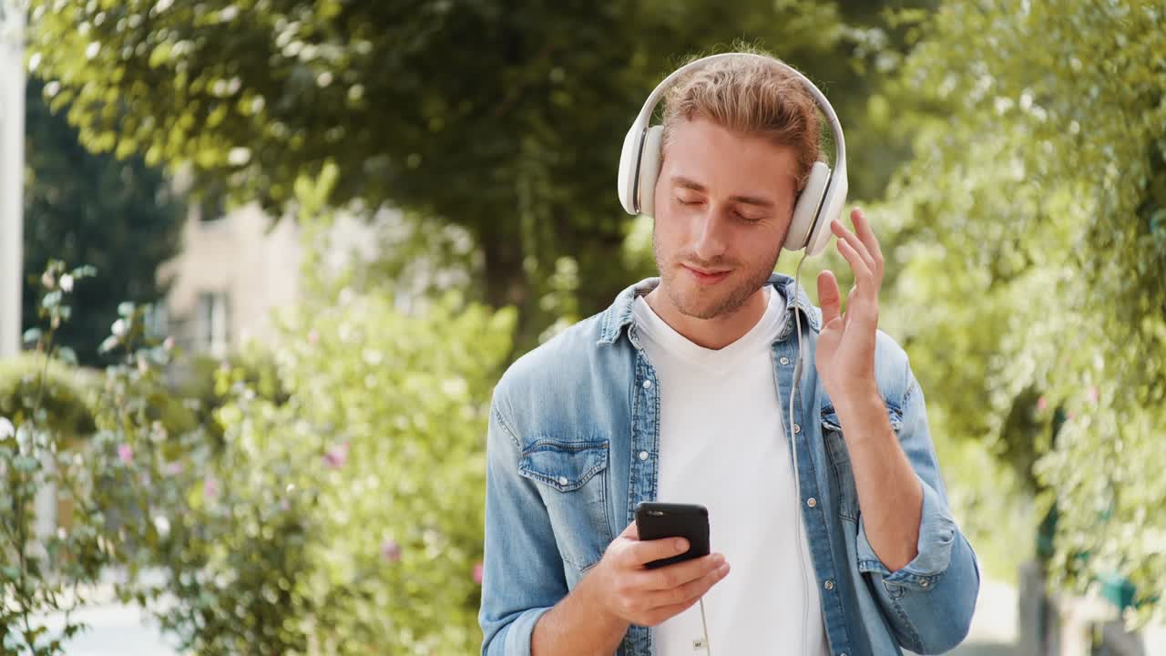 un hombre caucásico lindo está caminando en un día soleado con auriculares, navegando por internet. un hombre hipster guapo está caminando por el parque, escuchando música, teniendo un estado de ánimo agradable. buen día. estilo de vida en línea.