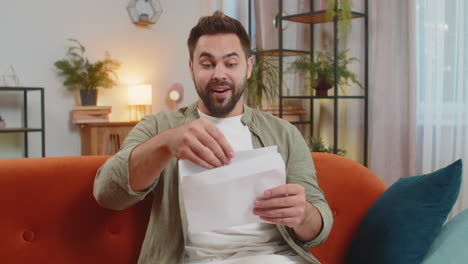excited young man opening envelope letter and reading good news sitting on sofa at home apartment