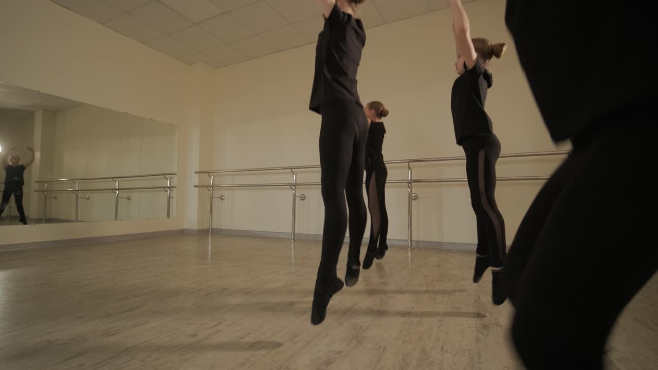 Un gruppo di giovani allievi di danza classica in abbigliamento da ballo nero che eseguono esercizi in un ampio studio di danza con pavimenti in legno e barre a muro. Espressioni concentrate e movimenti sincronizzati