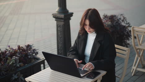 lady working on laptop outdoors in urban setting, wearing stylish black coat, with decorative black pillar nearby, blurred red brick building in background, and flower gently swaying in breeze