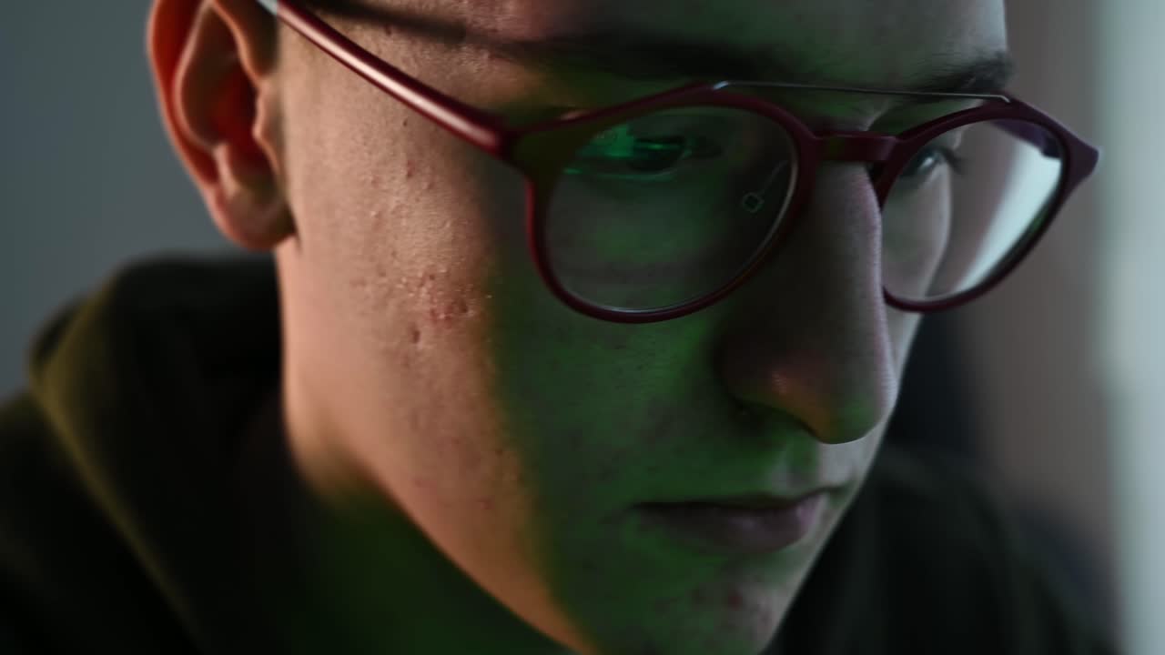 Young pimply man in red glasses writing codes on a green page in his computer. Concentrated programmer. Reflection in the glasses. Side view. Close up