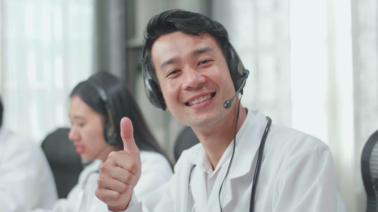 Close Up Of An Asian Man Doctor Wearing Headsets Working As Call Center Agent Sitting On The Chair Look Up, Smile, And Thumbs Up While His Colleagues Are Speaking And Typing During A Call At The Office