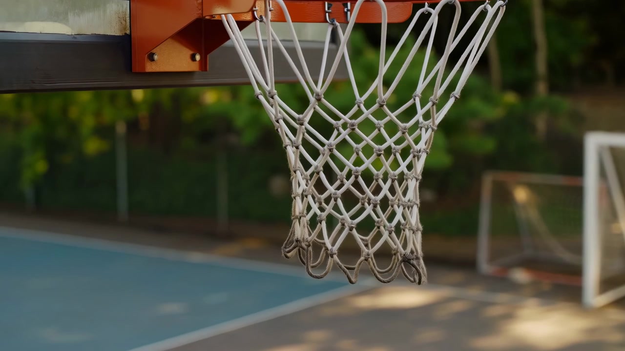 Vista laterale di un canestro da basket dipinto di arancione con rete di nylon bianca annodata che pende liberamente su un campo comunitario all'aperto, mostrando la texture del metallo invecchiato, luce solare soffusa, messa a fuoco superficiale e uno sport tranquillo.