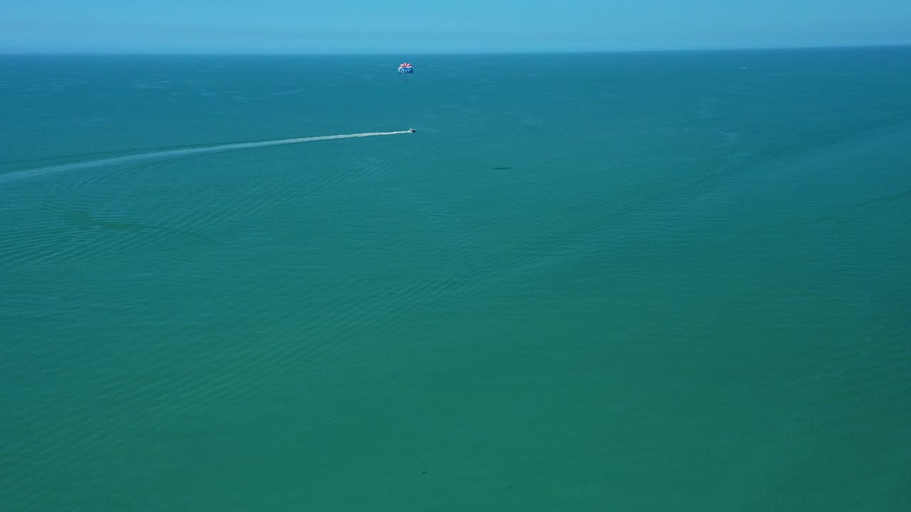 Un barco arrastra un parasail tipo paracaídas sobre aguas tranquilas y azules del océano, creando estelas, lo que ilustra una actividad deportiva acuática de alta adrenalina bajo cielos despejados y condiciones de mar abierto.