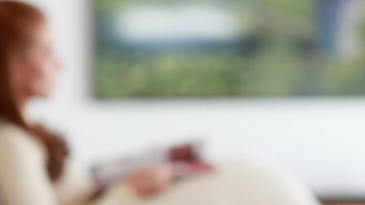 A woman sits on a sofa in a bright living room, holding a remote control and magazine, interacting with a large television screen. Soft natural lighting