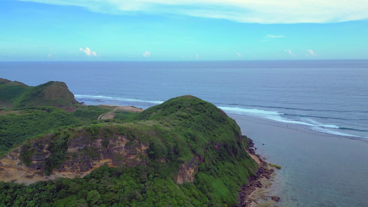 Tebing hijau rimbun menghadap samudra biru jernih dengan ombak kecil yang pecah, mengisyaratkan destinasi tropis. Pemandangan udara menakjubkan panorama terbalik terbang drone