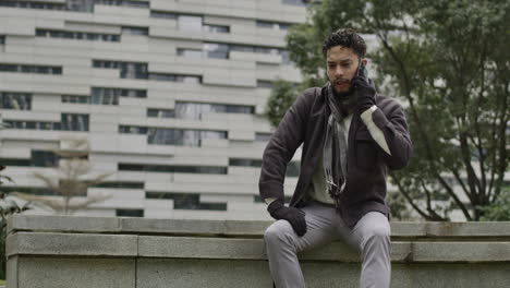 Caucasian-male-sitting-on-a-bench-of-a-residential-district-modern-area-while-talking-at-phone-in-slow-motion